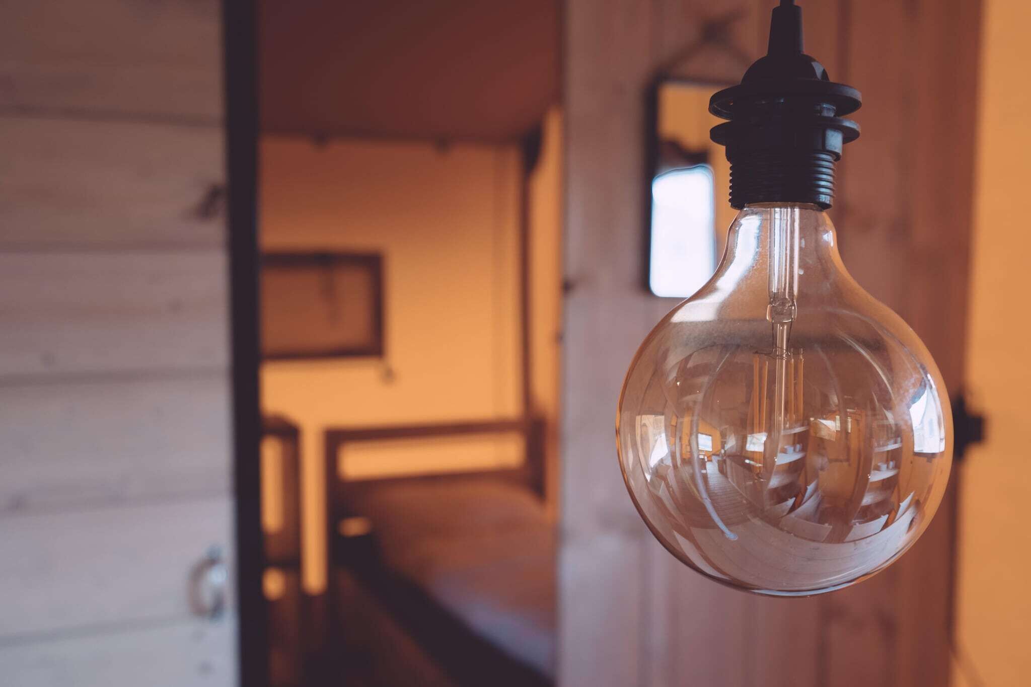 A clear glass light bulb hanging from a ceiling in a room with wooden walls and a visible staircase in the background.