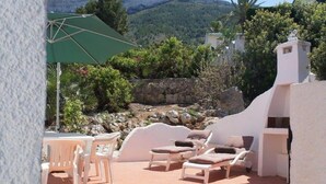 Een terras met witte stoelen en loungestoelen, een groene parasol, en uitzicht op een bergachtig landschap.