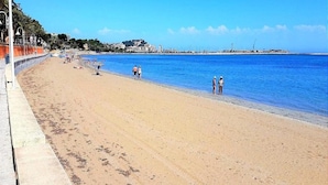 Een uitgestrekt zandstrand met een heldere blauwe zee, enkele mensen wandelen en een verre weergave van gebouwen en een schiereiland.