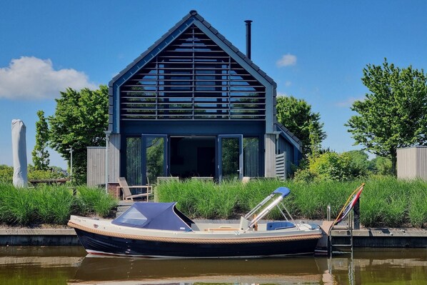 Ett modernt hus med ett sluttande tak och stora fönster, beläget vid en kanal där en båt ligger förtöjd framför.
