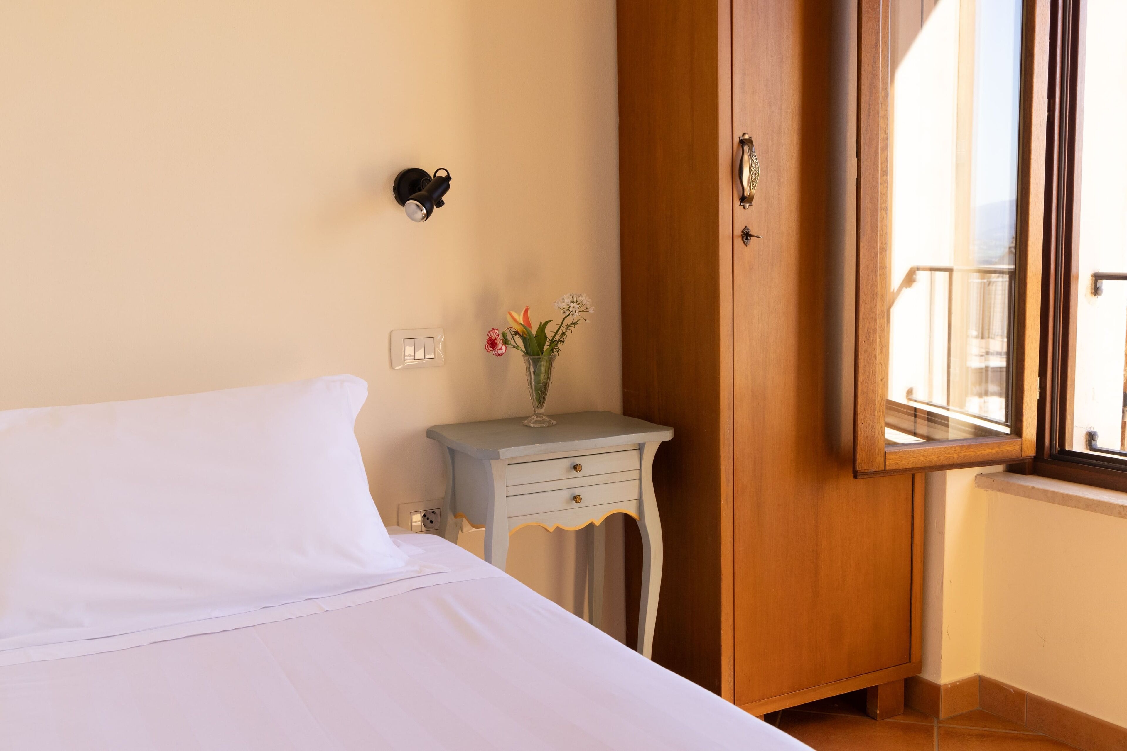 Habitación de hotel con una cama, una mesita de noche con un florero y una lámpara de pared, además de una puerta de madera con una ventana.