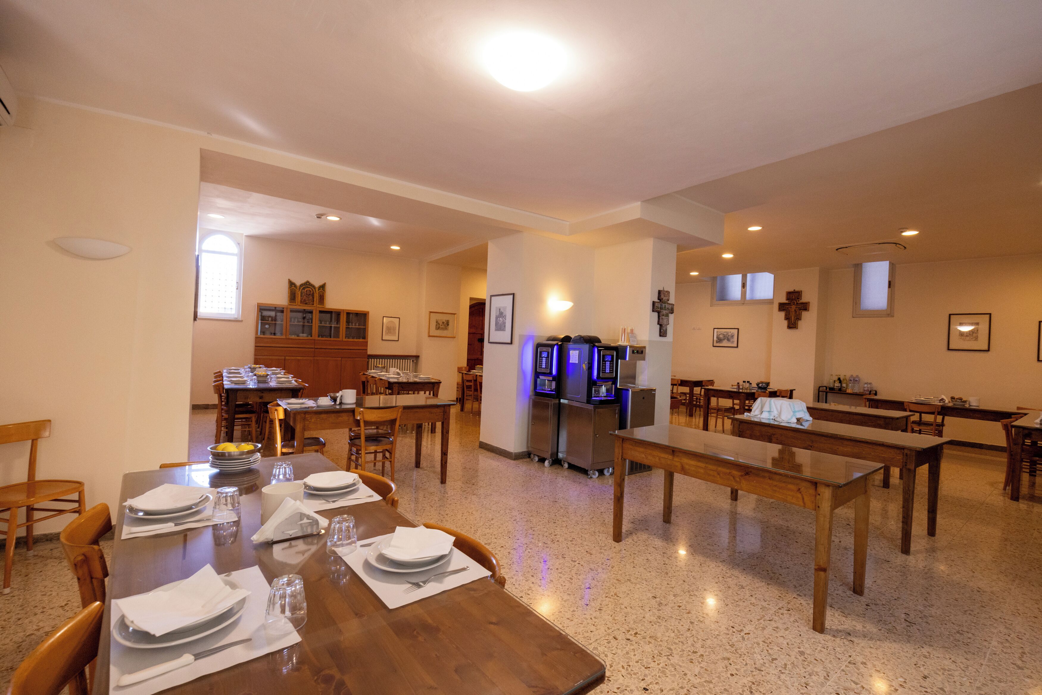 A dining area with wooden tables and chairs, a kitchen area with cabinets and a refrigerator, and a small altar with a cross.