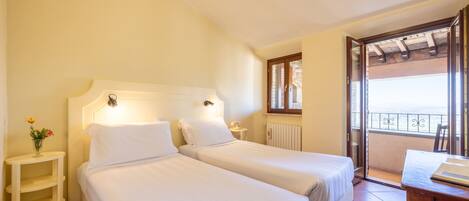 A hotel room with two single beds, a small bedside table with a lamp, a window with a view, and a balcony with a railing.