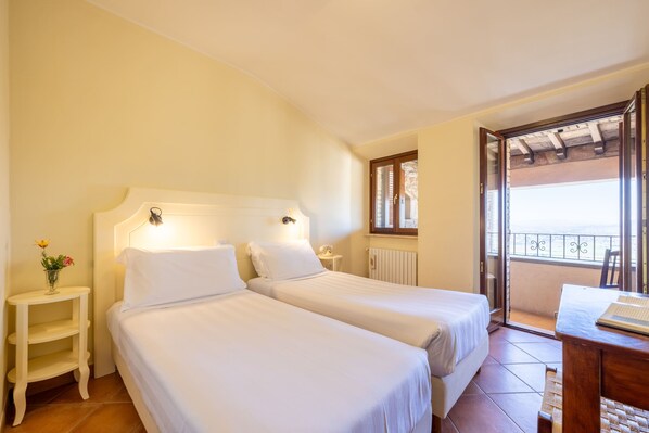 A hotel room with two single beds, a small bedside table with a lamp, a window with a view, and a balcony with a railing.