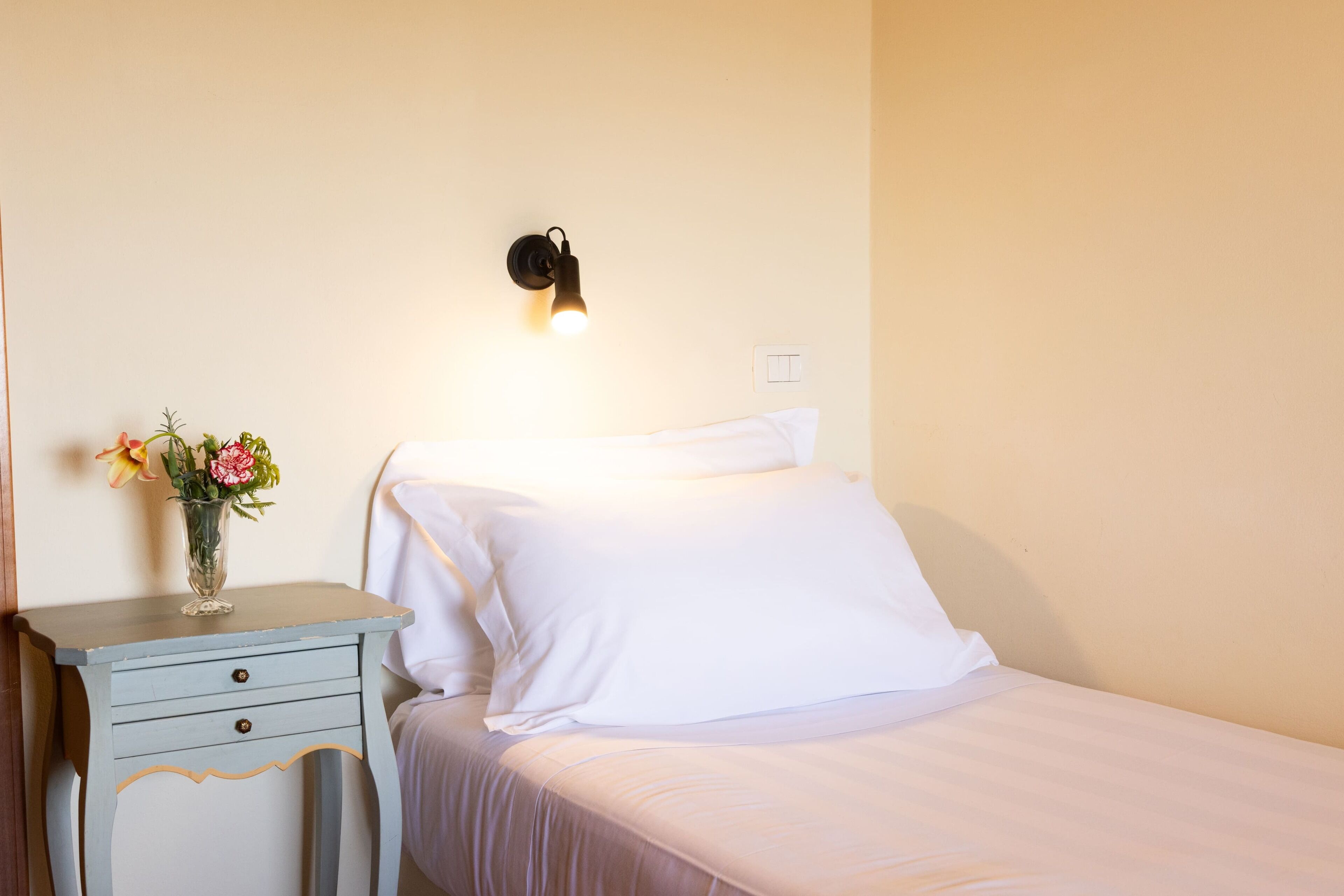 A bedroom with a bed, a nightstand with a vase of flowers, and a wall-mounted light.