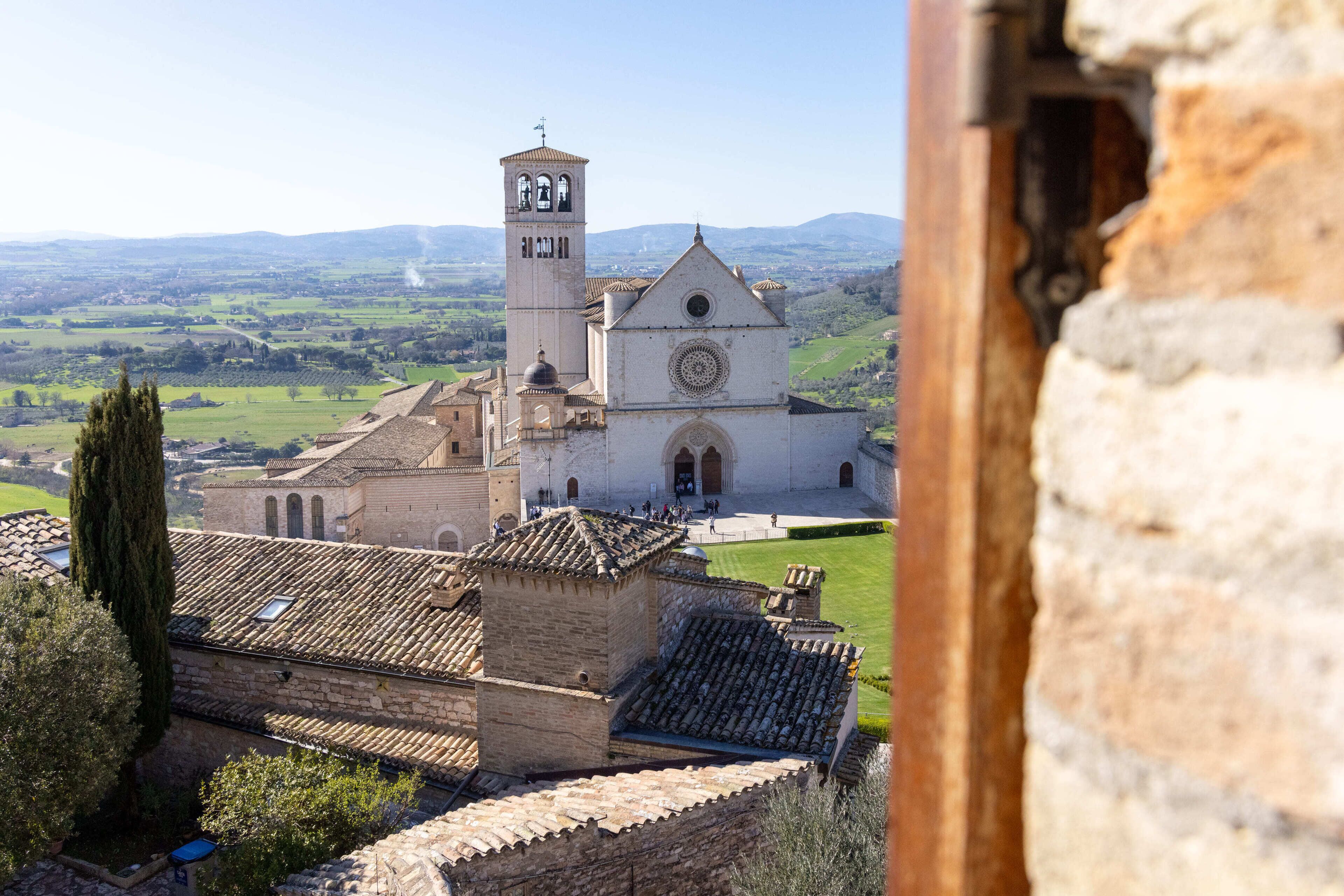 A historic building with a bell tower and a large circular window, surrounded by a lush landscape.