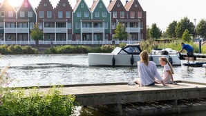 A wooden dock with two people sitting, a canal with boats, and colorful houses in the background. - Stylish Stay in Volendam (Volendam)