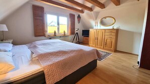 Un dormitorio con cama, vigas de madera, armario, escritorio y ventana con contraventanas.