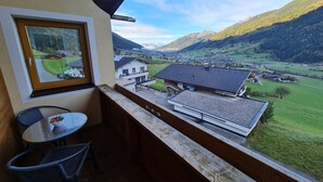 A balcony with a table and chairs, overlooking a valley with houses and mountains.