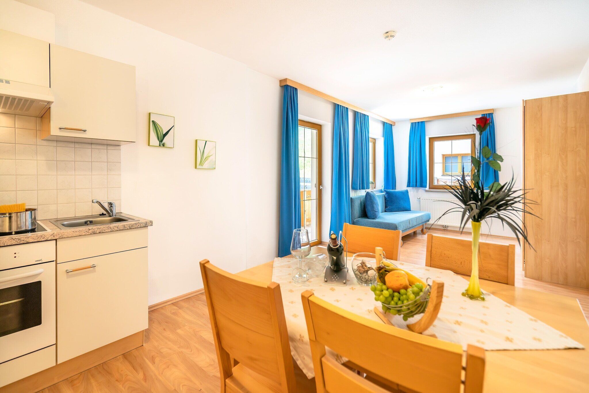 A compact kitchen with a sink and stove, a dining area with a table set for two, and a blue sofa in the living area.