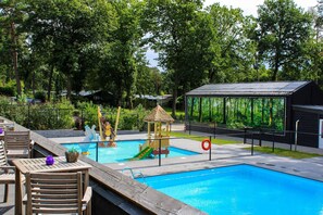 Une piscine avec un toboggan et un espace de jeux, entourée d’arbres et d’une terrasse clôturée avec des chaises en bois.