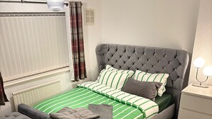 Un dormitorio con cama y cabecera capitonada, ropa de cama verde a rayas, un otoman gris y una mesita de noche con dos lámparas.