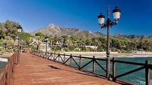 A wooden pier with metal railings and street lamps, leading to a beach with mountains in the background.