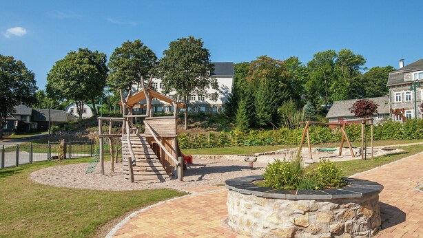 Une aire de jeux avec un toboggan en bois et une structure d’escalade, entourée d’une zone pavée et de verdure.