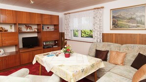Un salon chaleureux avec des meubles en bois, un téléviseur, une table à manger recouverte d’une nappe à motifs floraux et un canapé agrémenté de coussins.