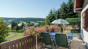 Un balcon avec une table et des chaises, donnant sur un paysage pittoresque avec des maisons et de la verdure.