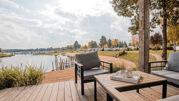 A wooden deck with outdoor seating, a view of a lake, and a row of RVs in the background.
