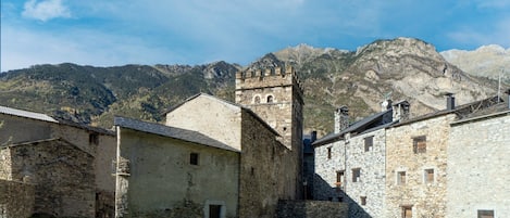Ein historisches Steinhaus mit einem Turm, umgeben von einem Kopfsteinpflasterweg und Rasenflächen, vor einer Bergkulisse.