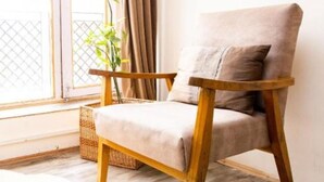 Una cadira amb coixí de color clar i estructura de fusta, col·locada a prop d'una finestra amb cortines, sobre un sostre de fusta i una catifa blanca i mullada.