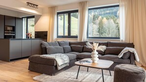 A modern living room with a sectional sofa, a coffee table, and a view of a forest through large windows. - Apartment with shared infinity pool (St. Martin am Tennengebirge)