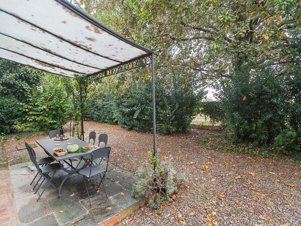 An outdoor dining area with a table and chairs under a shaded canopy, surrounded by trees and a gravel path.