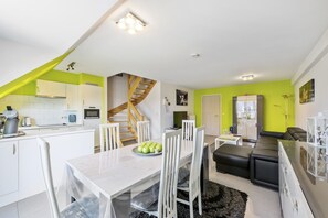 A modern kitchen with white cabinets, a dining area with a white table and chairs, a staircase, and a living area with a black sofa and a TV. - Nice duplex located in the market square (Blankenberge)