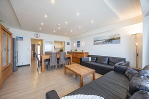 A spacious living area with a wooden coffee table, a black leather sofa, and a dining table with chairs. A kitchen is visible in the background, and there are wall-mounted lights.
