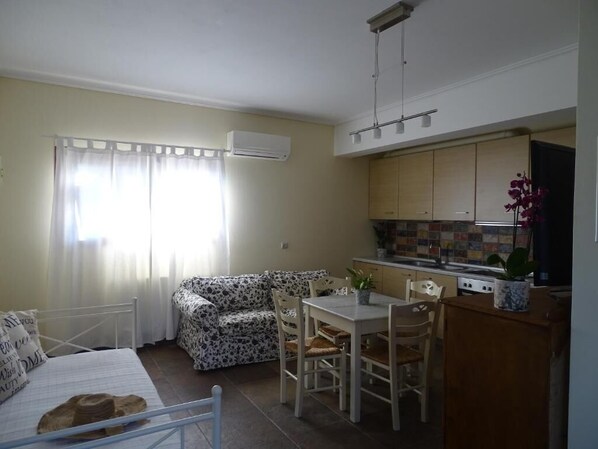 Ein kompakter Wohnraum mit Bett, Sofa, Esstisch mit Stühlen, Küchenzeile mit Schränken und einem Fenster mit Vorhängen.
