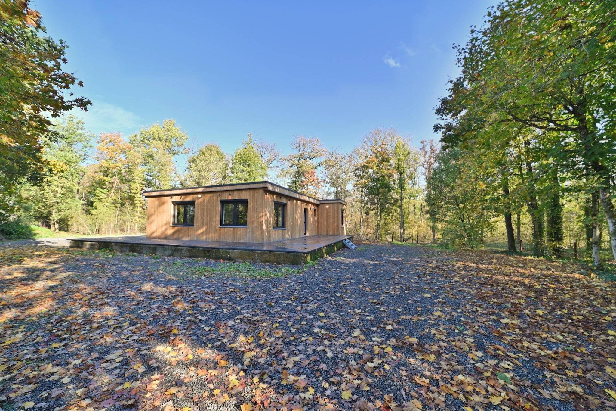 A modern wooden cabin with a deck, surrounded by a forest with autumn leaves.