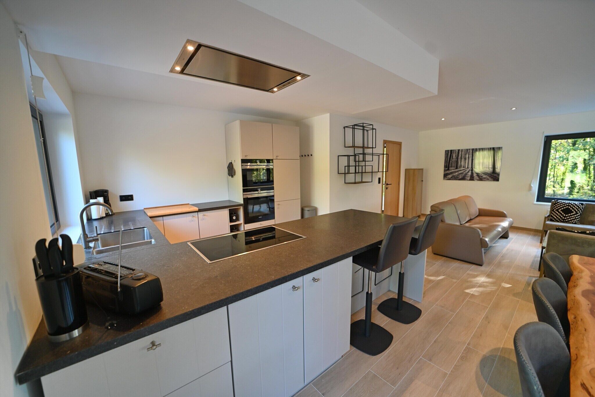 Een moderne keuken met een centraal eiland, witte kasten en een ingebouwde oven. Er is een zithoek met een bank en stoelen.
