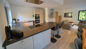 Une cuisine moderne dotée d’un îlot central, de meubles blancs et d’un four encastré. On y trouve également un coin repas avec un canapé et des chaises.