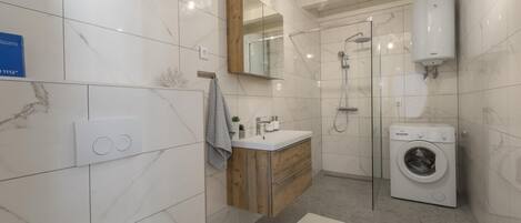 Une salle de bain moderne avec un mur recouvert de carreaux de marbre, un meuble-lavabo en bois, une cabine de douche en verre et un lave-linge encastré.