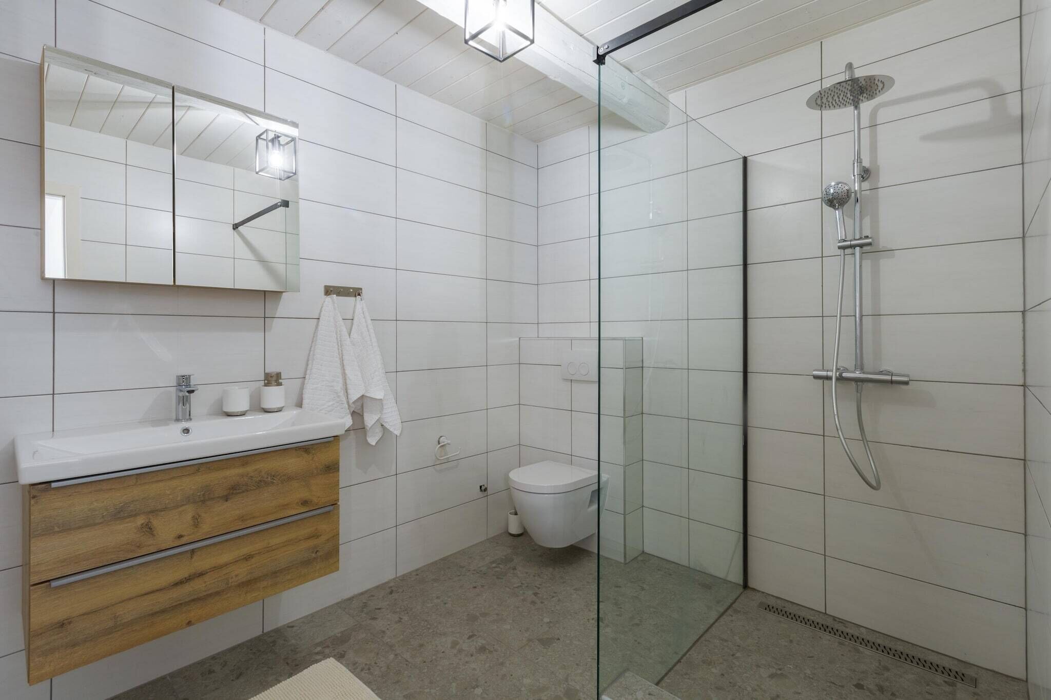 Moderni kylpyhuone, jossa on lasiseinäinen suihkukaappi, puinen allaskaappi peilillä sekä wc-istuin.