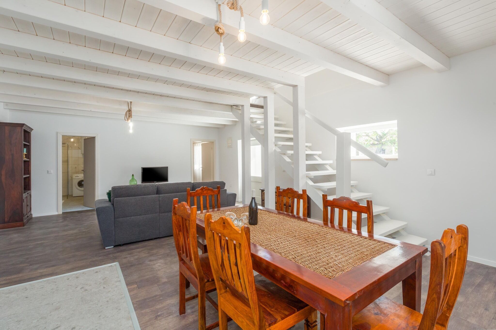 Un vaste espace de vie comprenant une table à manger en bois, des chaises et un canapé. Un escalier mène à l’étage supérieur.