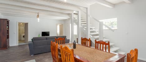 Un vaste espace de vie comprenant une table à manger en bois, des chaises et un canapé. Un escalier mène à l’étage supérieur.