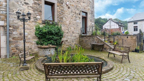 A stone building with a courtyard featuring a wooden bench, a small garden, and a staircase leading to an upper level.