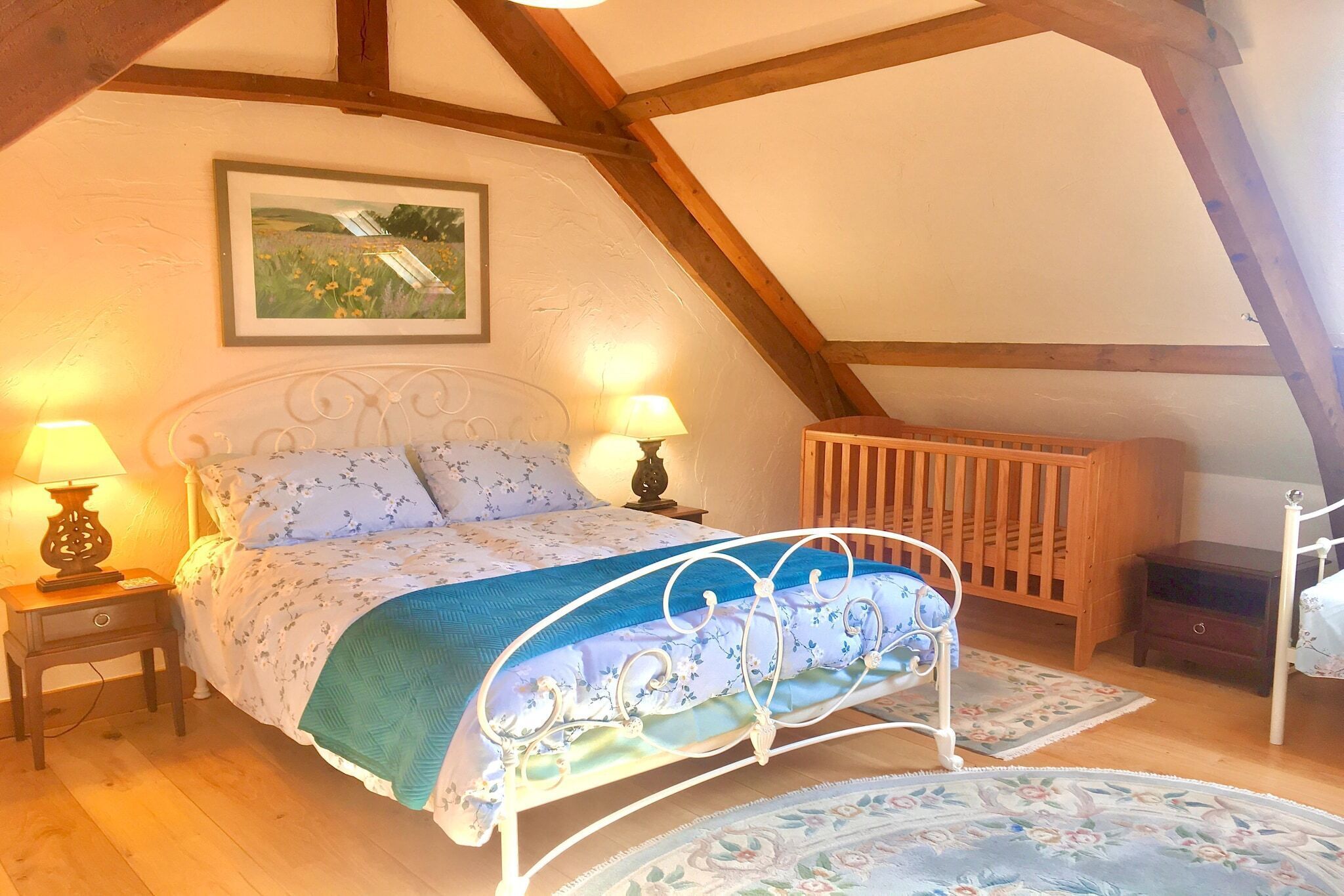 A bedroom with a bed, two bedside tables with lamps, a crib, a wooden bench, and a framed picture on the wall.