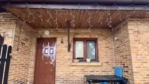 A covered outdoor area with a brick wall, a wooden door, a window with a view, a table with a blue chair, and a hanging light fixture. - Vector Home (Warsaw)