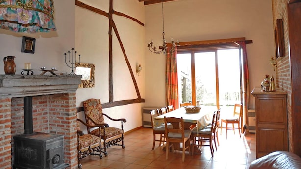 Uma sala de jantar com lareira de tijolos, vigas de madeira, um candelabro e vista através de uma grande janela.