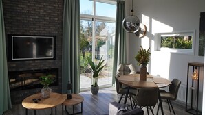 A modern living room with a fireplace, a dining area, and large windows offering a view of the outdoors. - Forest-View Tiny House (Hoenderloo)
