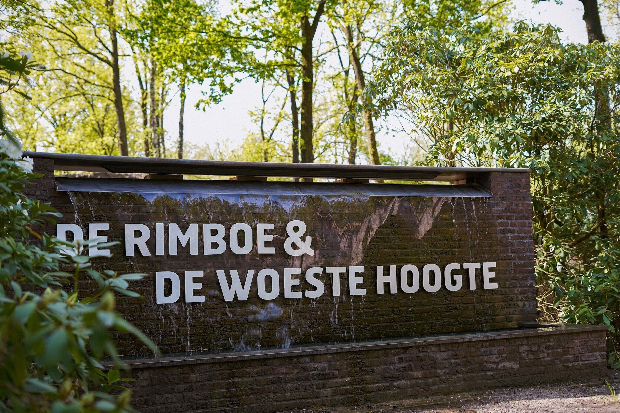 A sign with the text "DE RIMBOE & DE WOESTE HOOGTE" in a natural setting with trees and greenery.