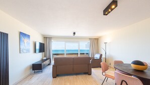 A modern living room with a sofa, a dining table with chairs, a TV, and a large window with a view of the sea.