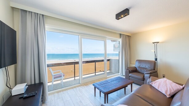 A room with a large sliding glass door leading to a balcony with a view of the beach. There is a sofa, a chair, and a small table with a remote control on it.