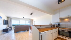 A modern kitchen with white cabinets, a wooden countertop, and an oven. There is a dining area with a brown sofa and a TV on the left.