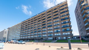 A multi-story residential building with balconies, a parking area, and a clear sky.