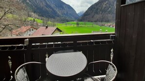 A balcony with two chairs and a table, overlooking a valley with green fields and houses, with mountains in the background.