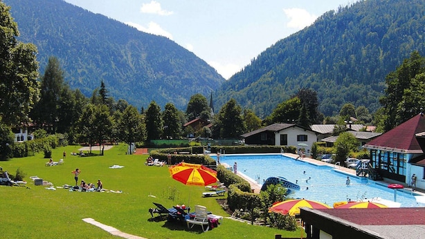 Un espace piscine doté de chaises longues et de parasols, entouré d’une pelouse verdoyante et avec des montagnes en toile de fond.