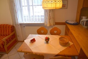 Une pièce chaleureuse avec une table en bois, deux chaises en forme de cœur, une petite lampe de table, un radiateur, une fenêtre avec des rideaux et un canapé.