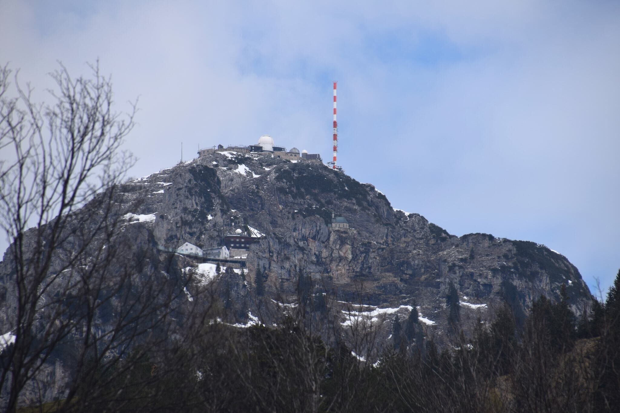 A mountain with a building on top, a red and white tower, and snow on the peak.