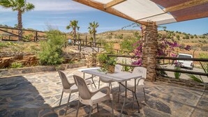 Een terras buiten met stenen vloer, een tafel met stoelen en uitzicht op een glooiend landschap met palmboompjes en paarse bloemen.